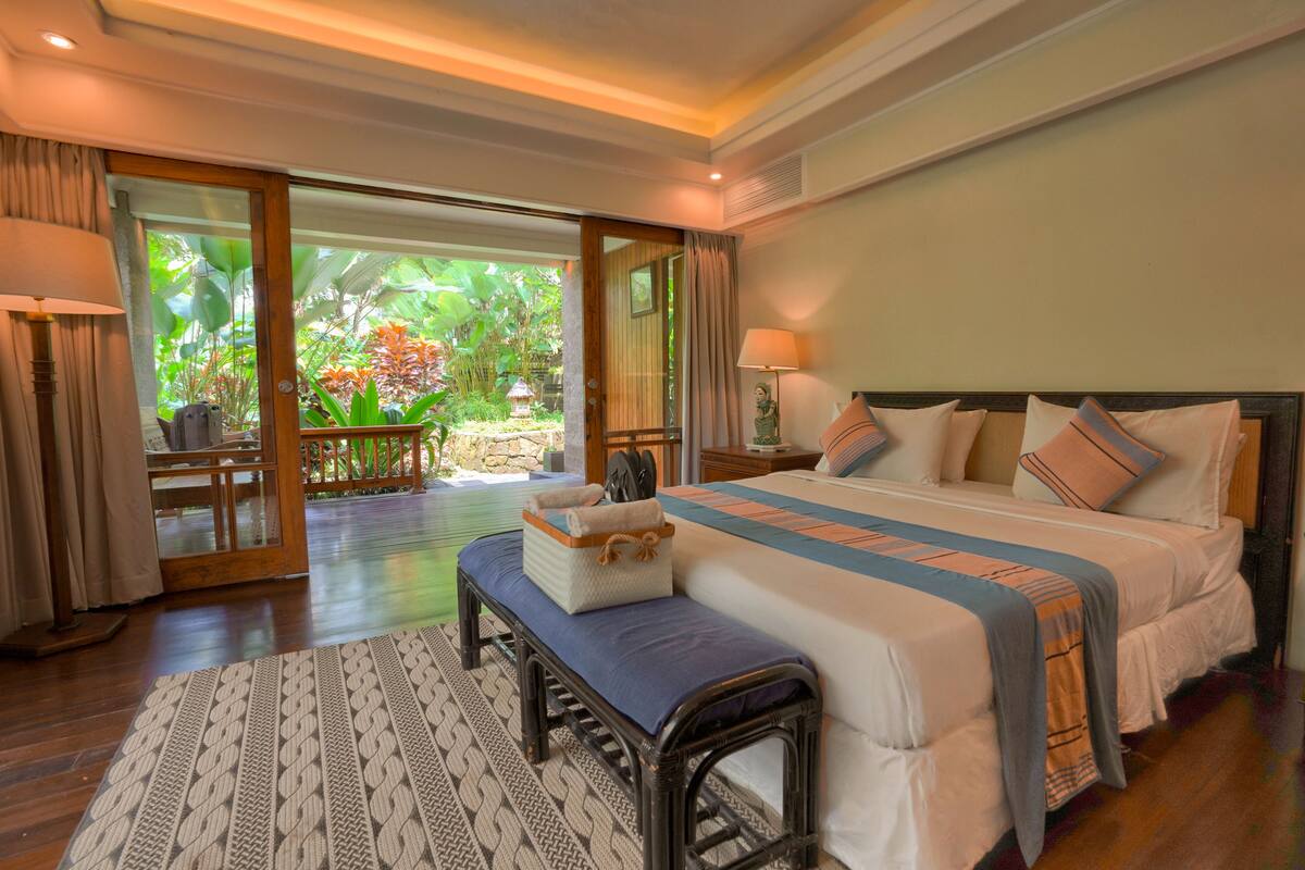 A spacious bedroom with a large bed, a bench, and a view of a lush garden through glass doors.