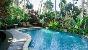 A swimming pool with a wooden deck, surrounded by lush tropical plants and palm trees. - Umah Lawas Ubud (Ubud)
