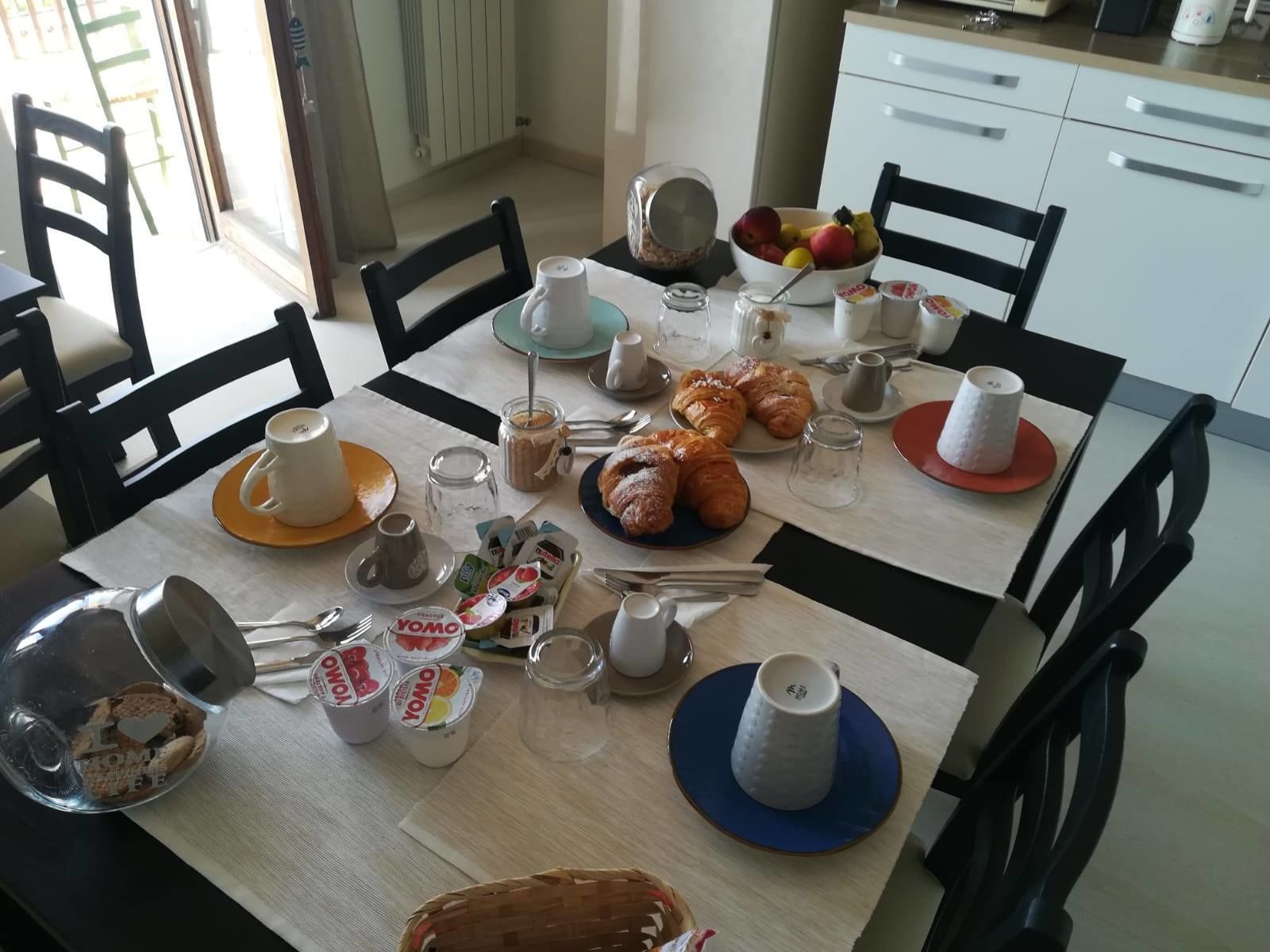 A breakfast table set with croissants, pastries, a fruit bowl, coffee cups, and condiments.
