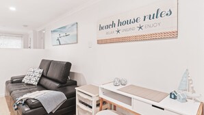 Un soggiorno moderno con un divano in pelle nera, un tavolino da caffè bianco e una targa a parete con la scritta "beach house rules", una stella marina e le parole “RELAX UNWIND ENJOY”.