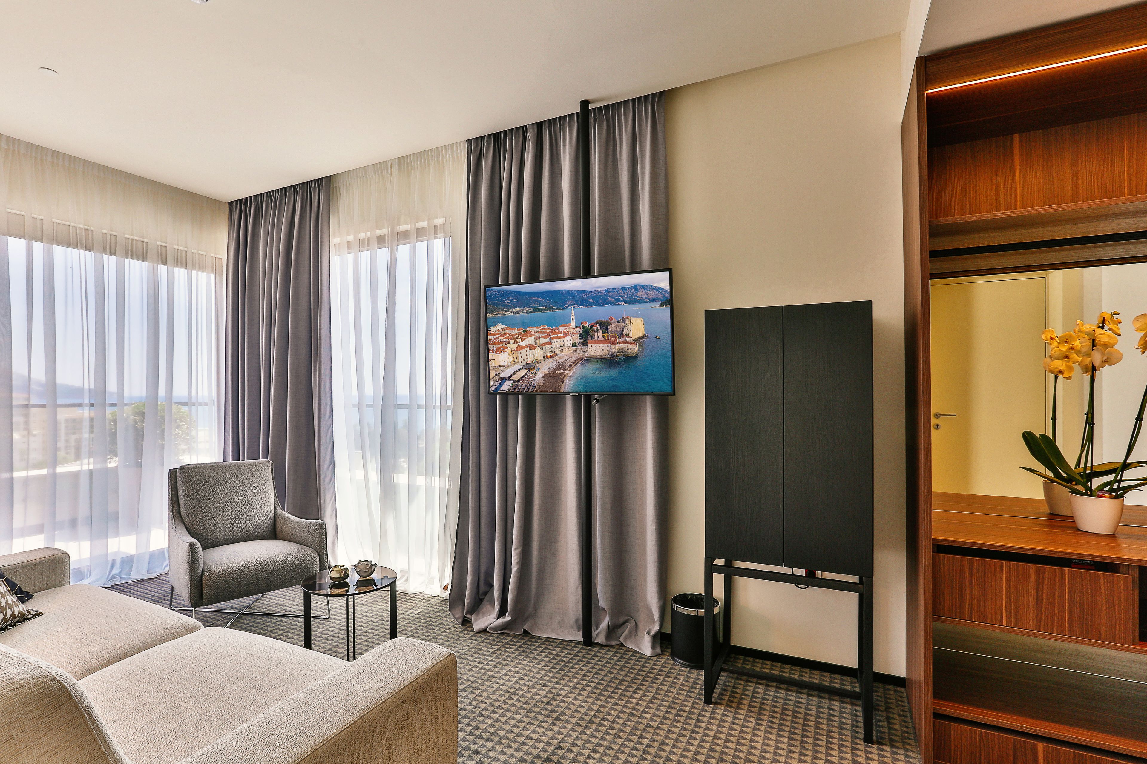 A modern hotel room with a large window, a flat-screen TV, a comfortable sofa, and a small table with decorative items.