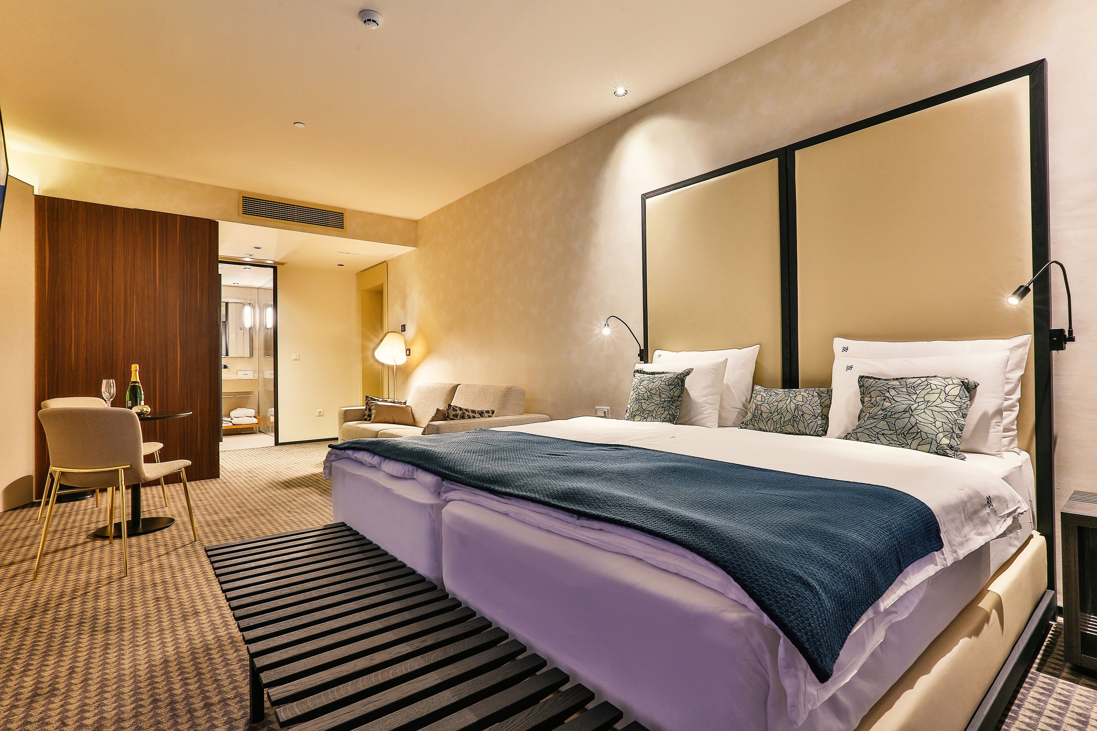 Habitación de hotel con una cama grande, un escritorio con silla, una mesita de noche con lámpara y un espejo grande en la pared.