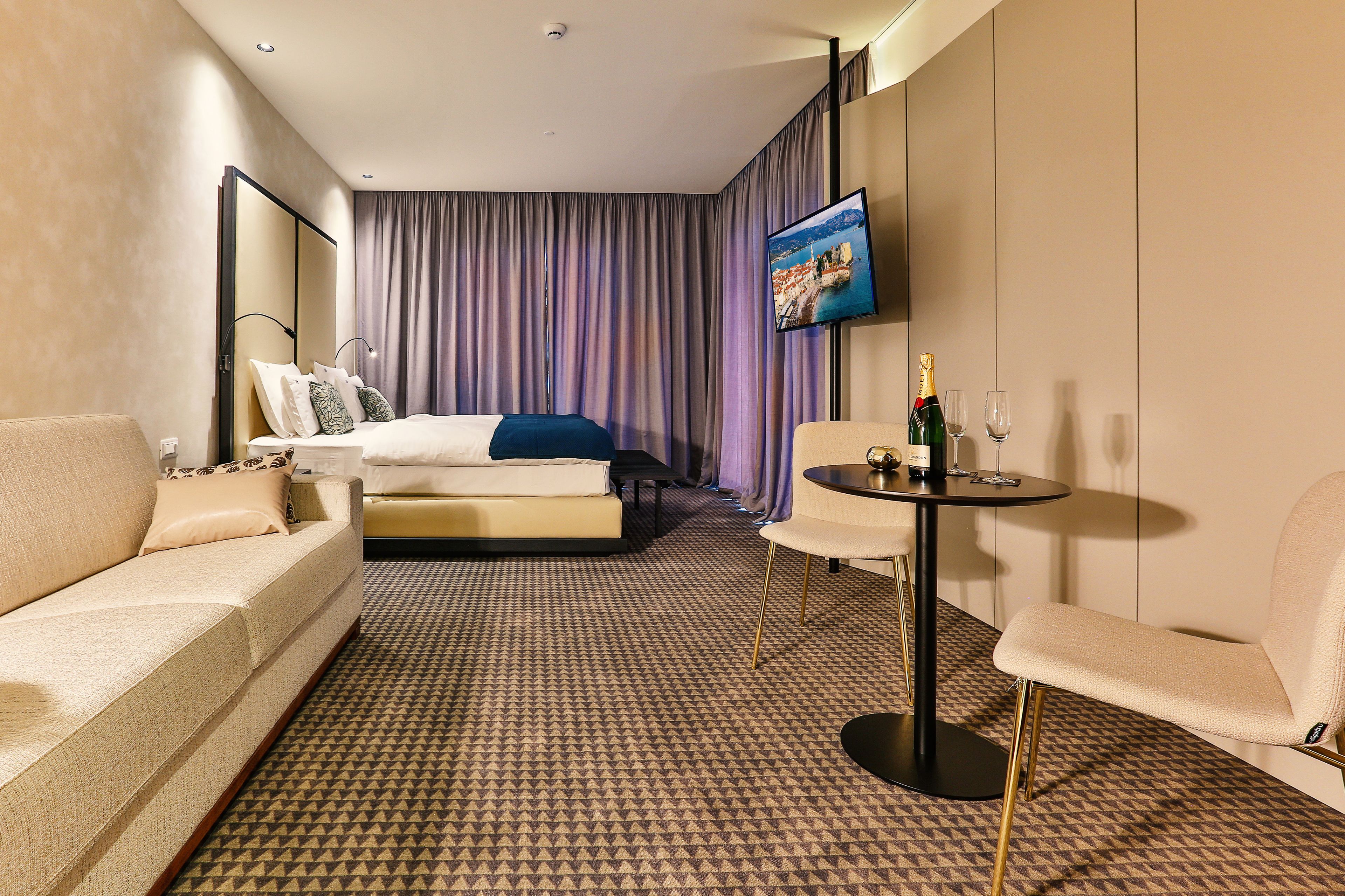 Una habitación de hotel con una cama, un sofá, una mesita con una botella y un vaso, y un televisor en la pared.