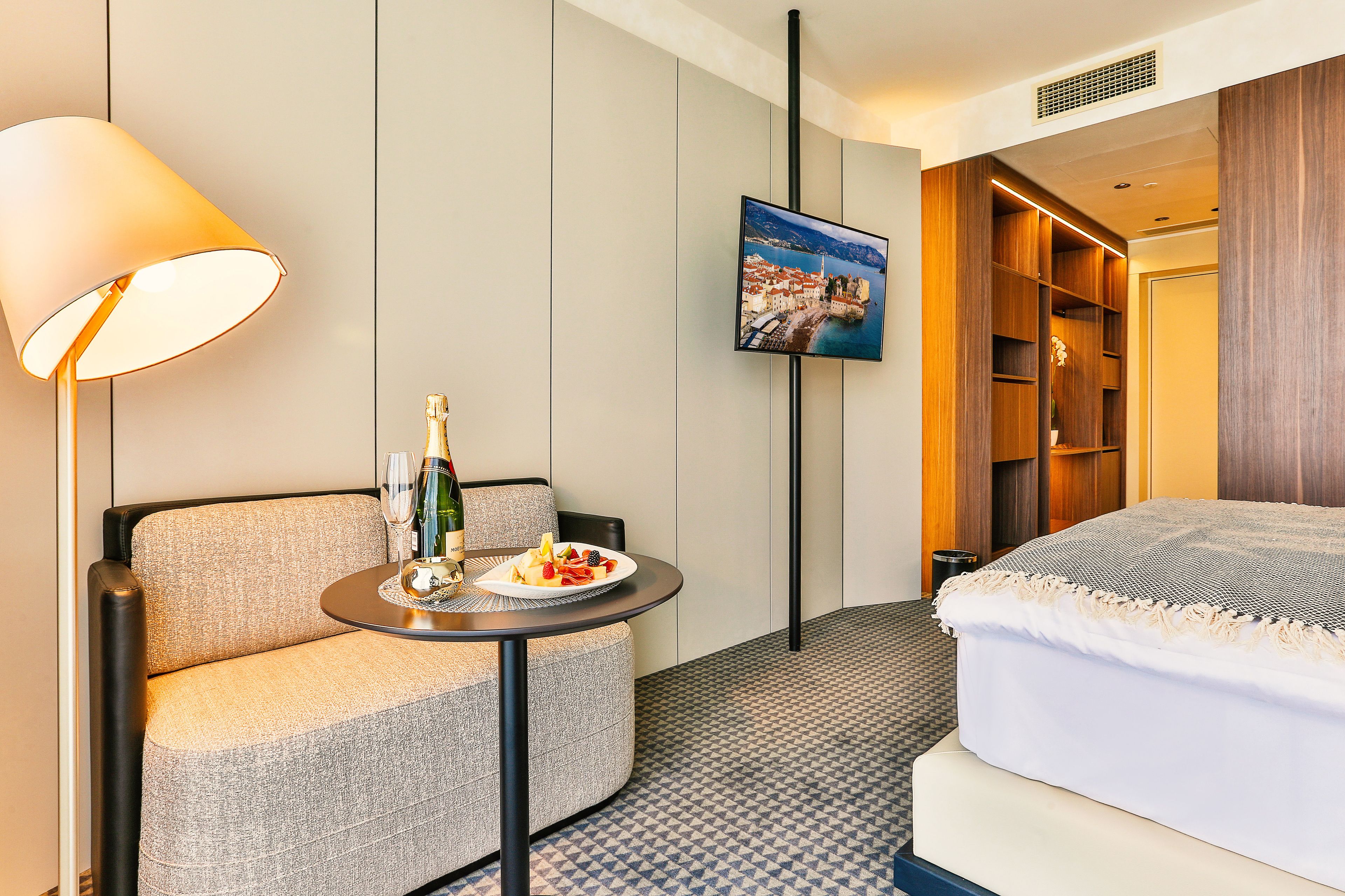 A hotel room with a sofa, a small table with a bottle and glasses, a bed with a white comforter, a TV on the wall, and a wooden wardrobe.