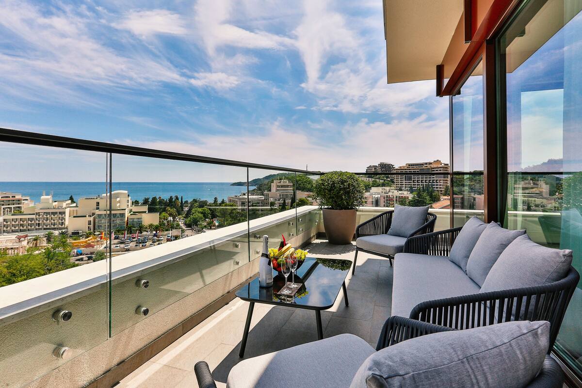 A balcony with a glass railing, a sofa, a table with a bottle and flowers, and a city view.