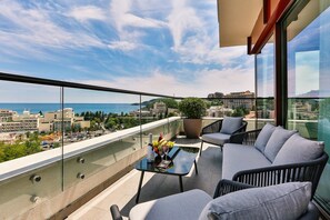 A balcony with a glass railing, a sofa, a table with a bottle and flowers, and a city view. - Hotel Kalos (Boreti)