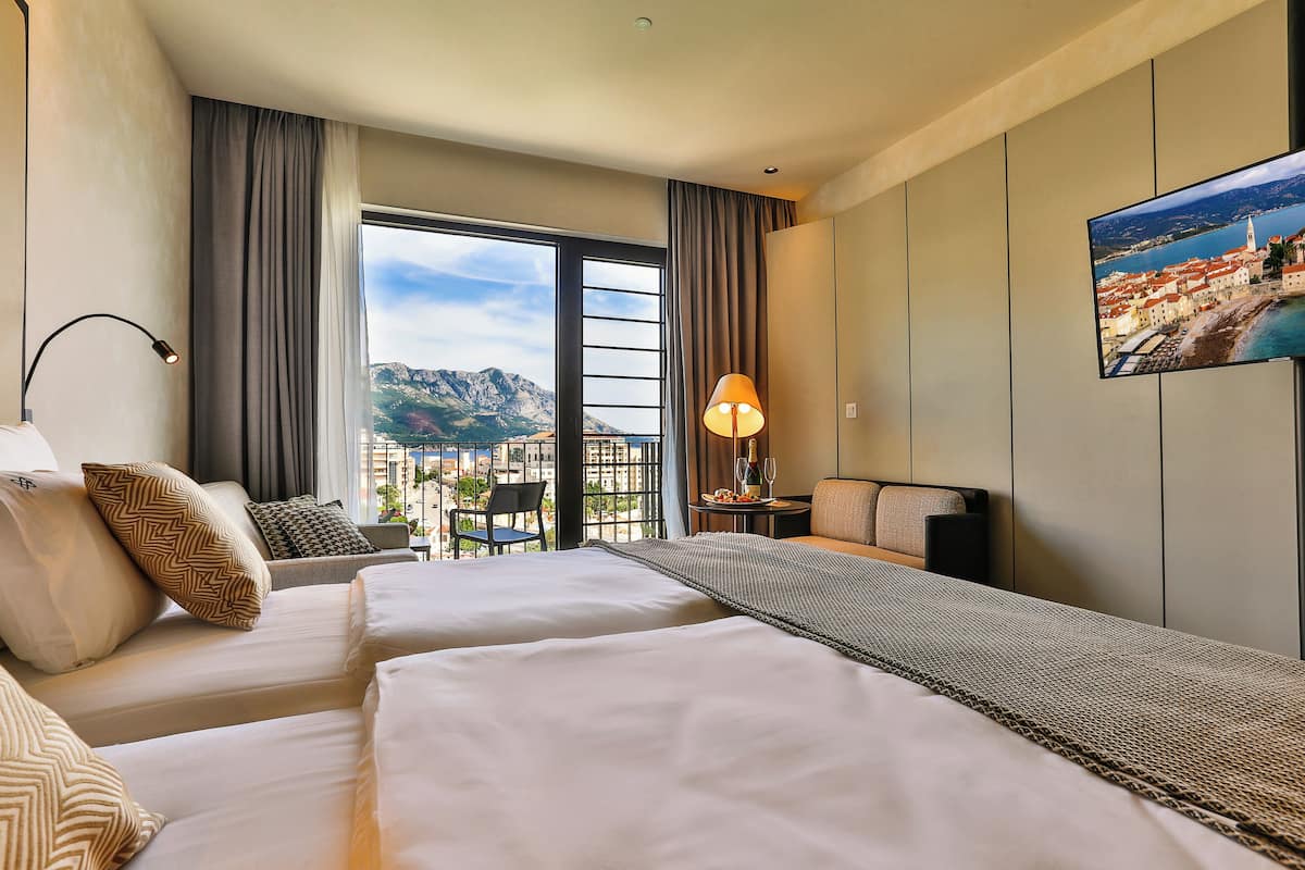 A hotel room with two beds, a view of a cityscape, a flat-screen TV, and a balcony with a table and chair.