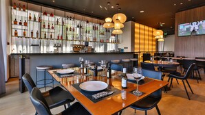 Un restaurante con zona de bar, mesas de madera, sillas negras y una pantalla de televisión.