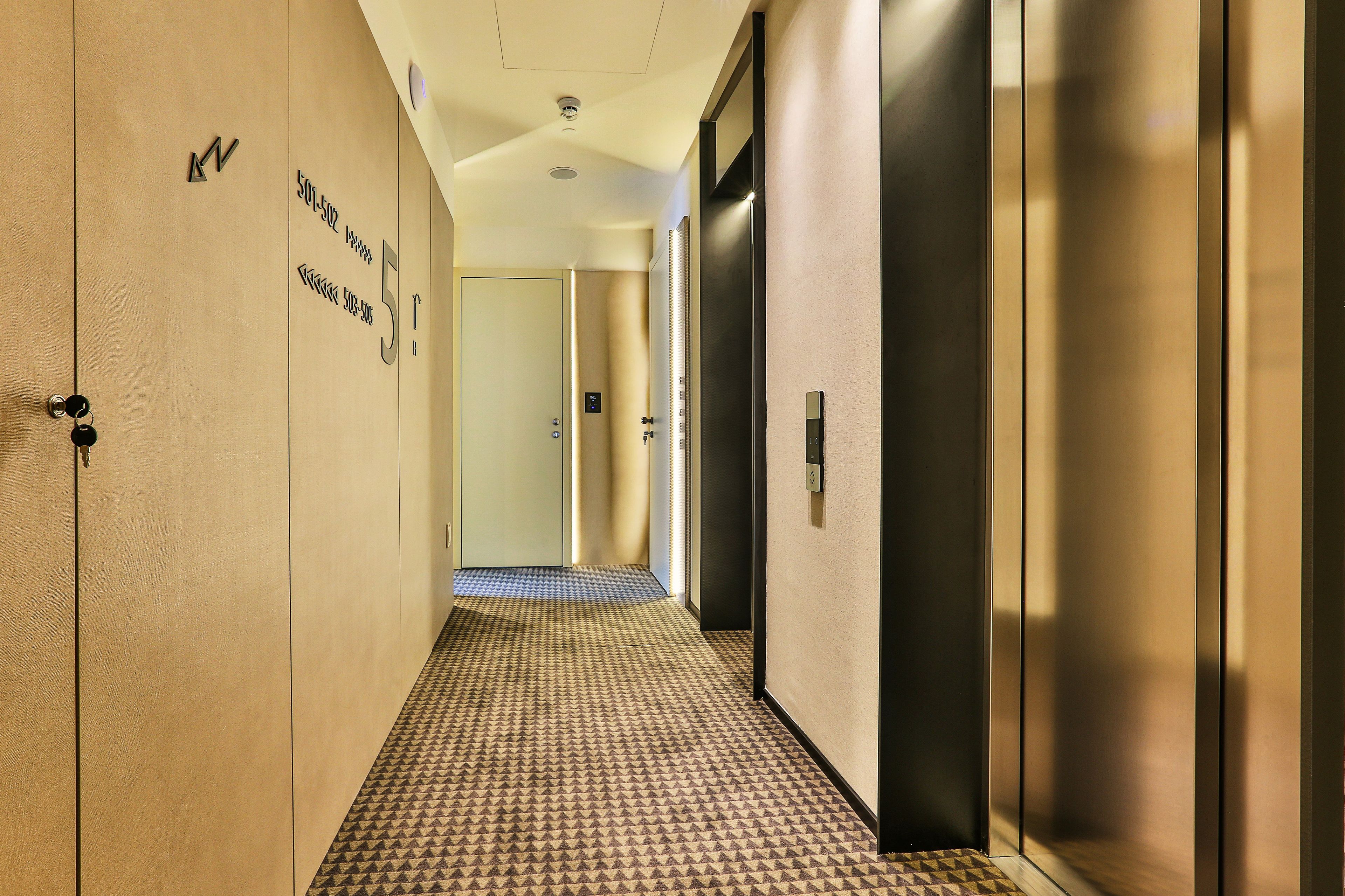 A hotel hallway with a patterned carpet, a door with a number, and an elevator.