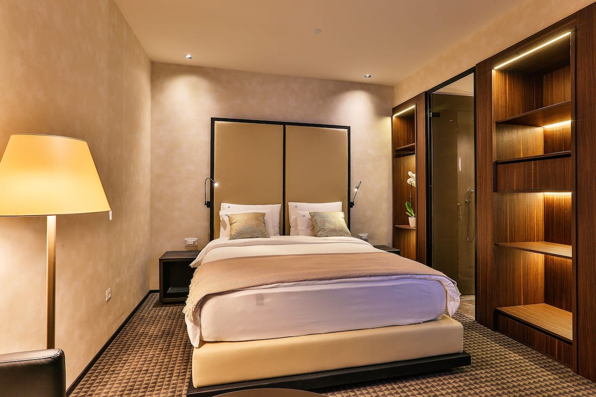A modern hotel room with a large bed, a bedside table, a lamp, a mirror, and built-in wooden shelving.