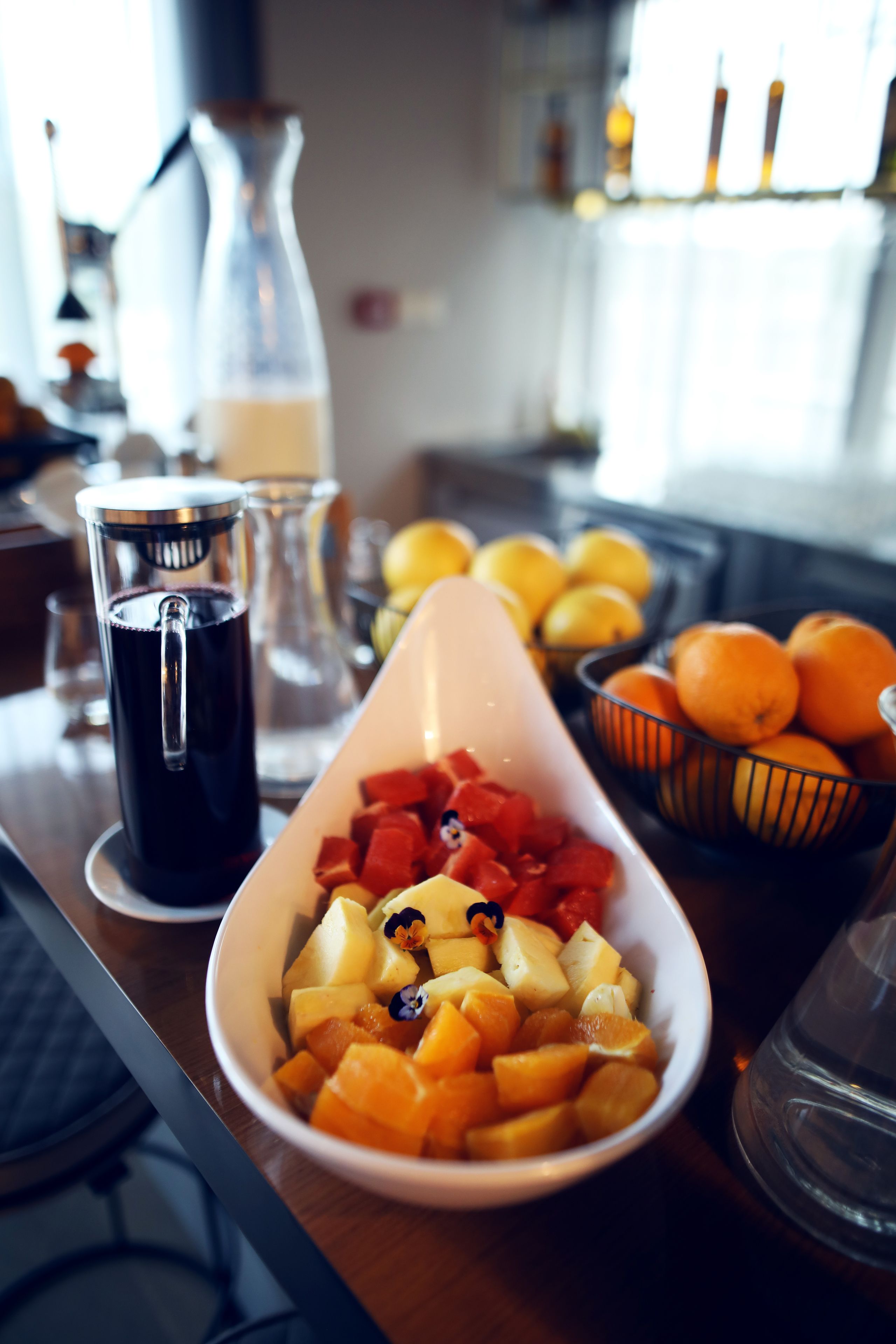 Uma mesa de café da manhã com uma fruteira, um jarro de suco e uma tigela de laranjas.