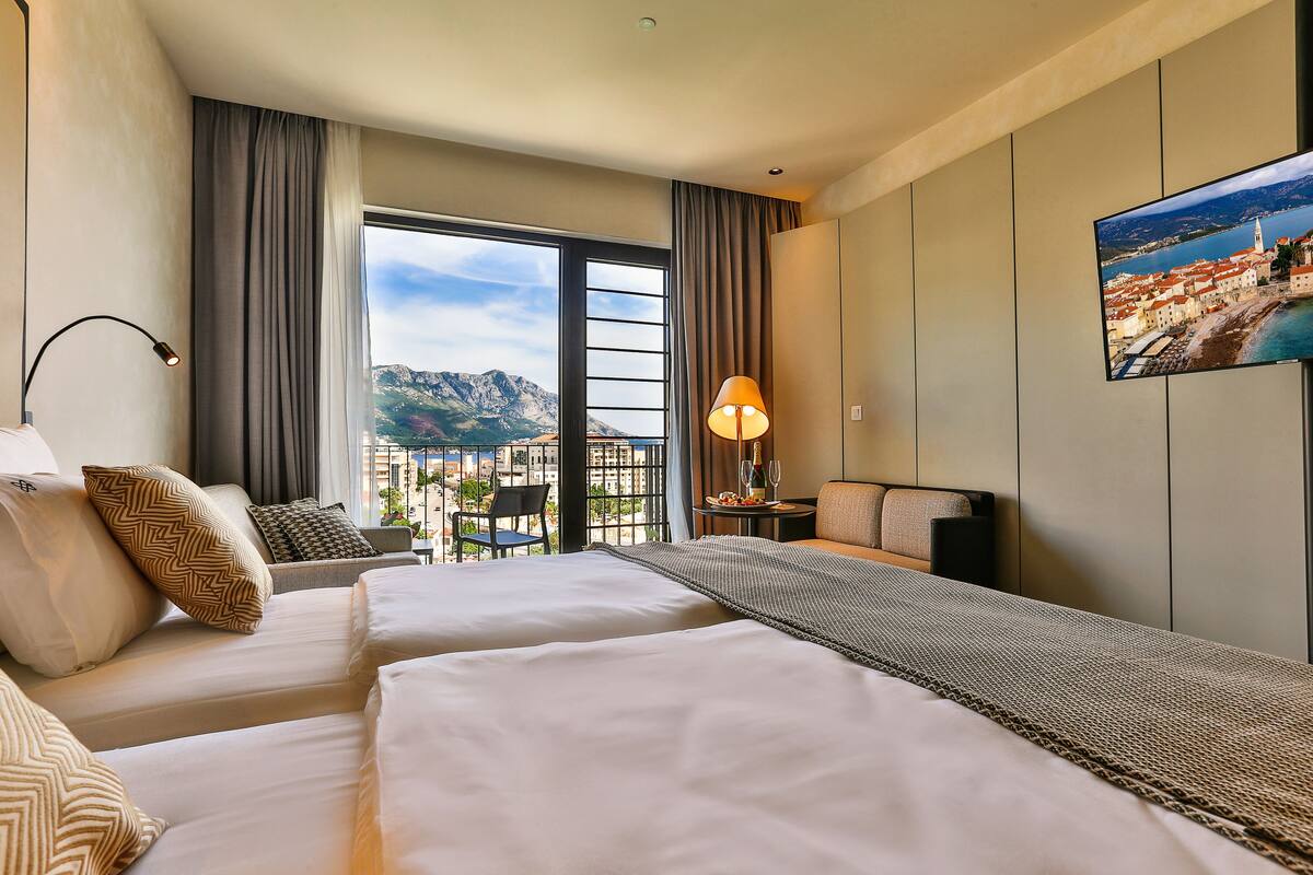 A hotel room with two beds, a view of a cityscape, a flat-screen TV, and a balcony with a table and chair.