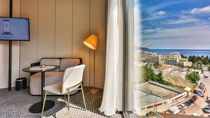 Habitación de hotel con sofá, sillón, televisión, lámpara de pie y vistas a la ciudad y al mar.