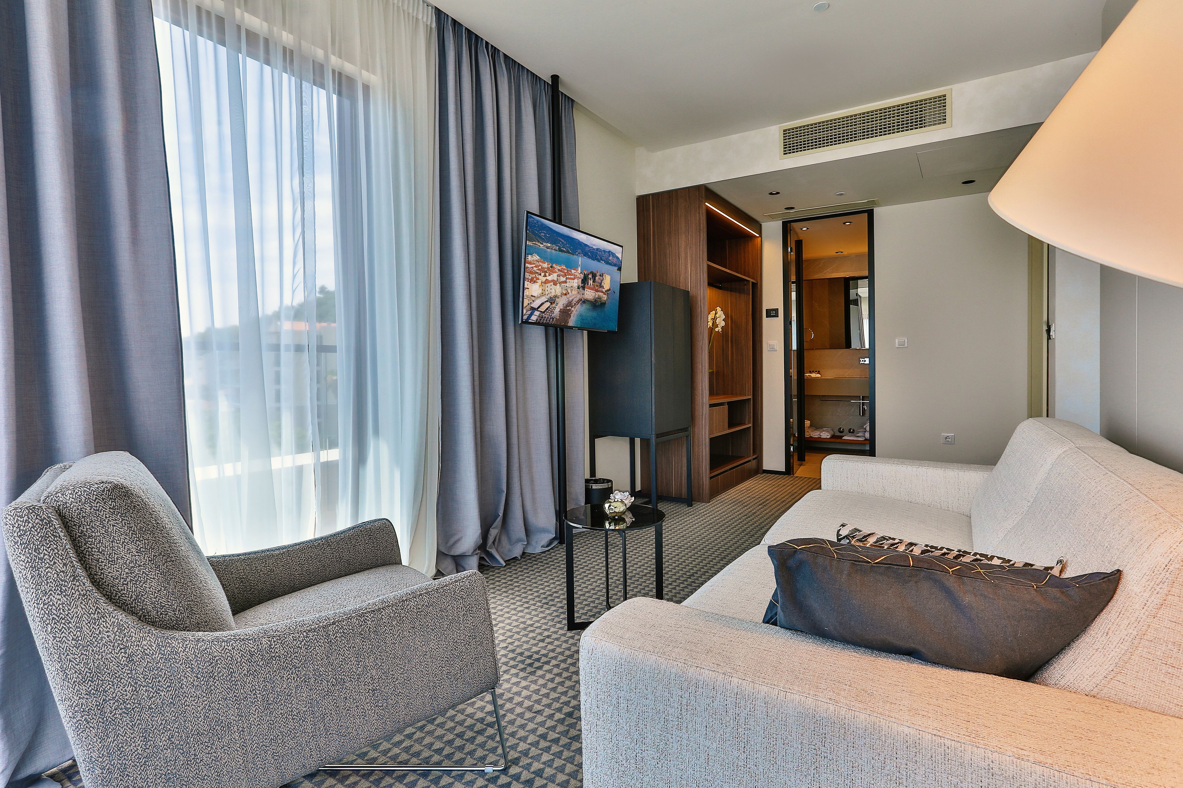 A modern hotel room with a sofa, armchair, and a flat-screen TV mounted on the wall.