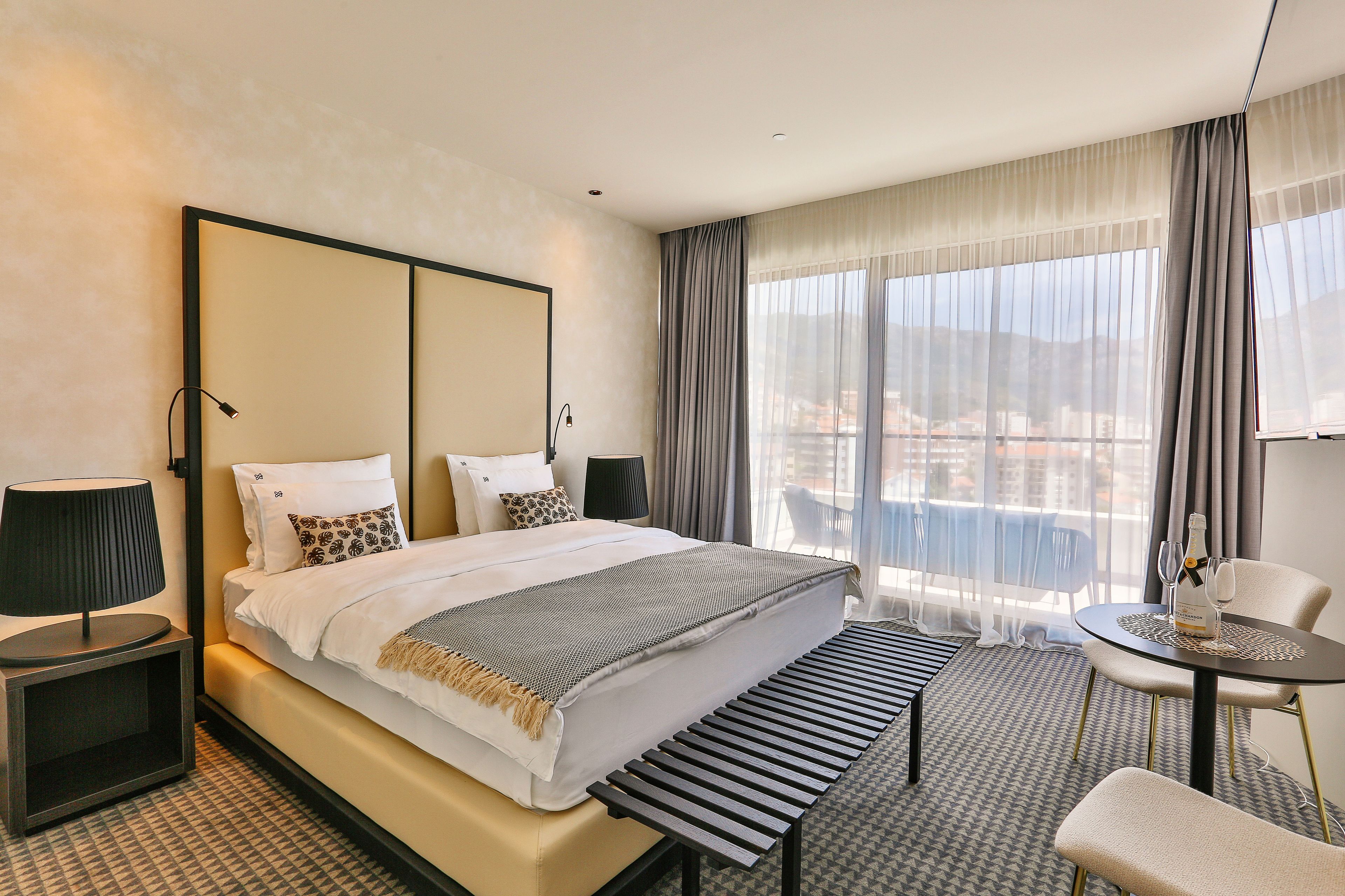 A hotel room with a large bed, bedside tables, a small table with a bottle and glasses, a chair, and a view of the city through large windows.