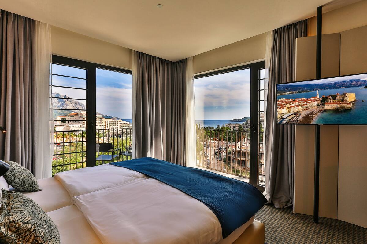 A hotel room with a large bed, a TV mounted on the wall, and a balcony with a view of the city and mountains.