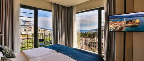 Habitación de hotel con una cama grande, televisión montada en la pared y balcón con vistas a la ciudad y las montañas.
