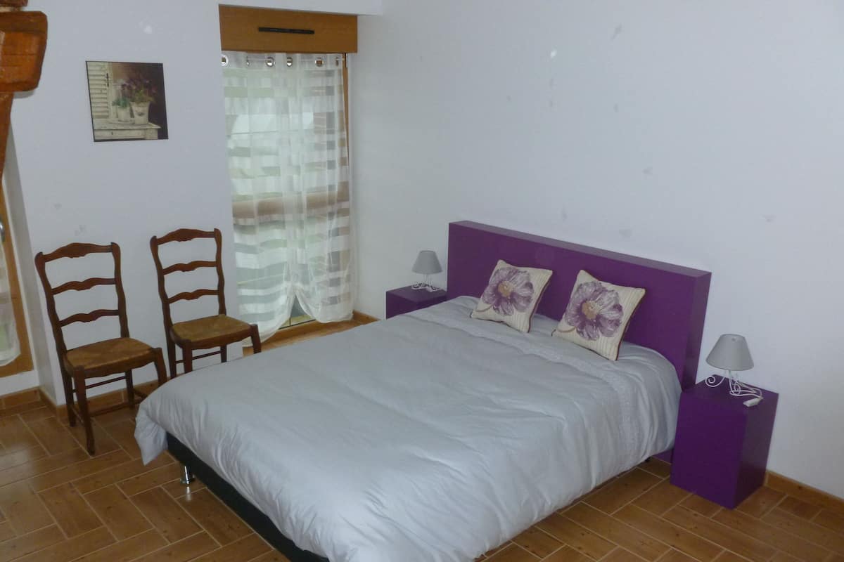 A bedroom with a bed, two nightstands, a chair, a window with curtains, and a wall-mounted picture.