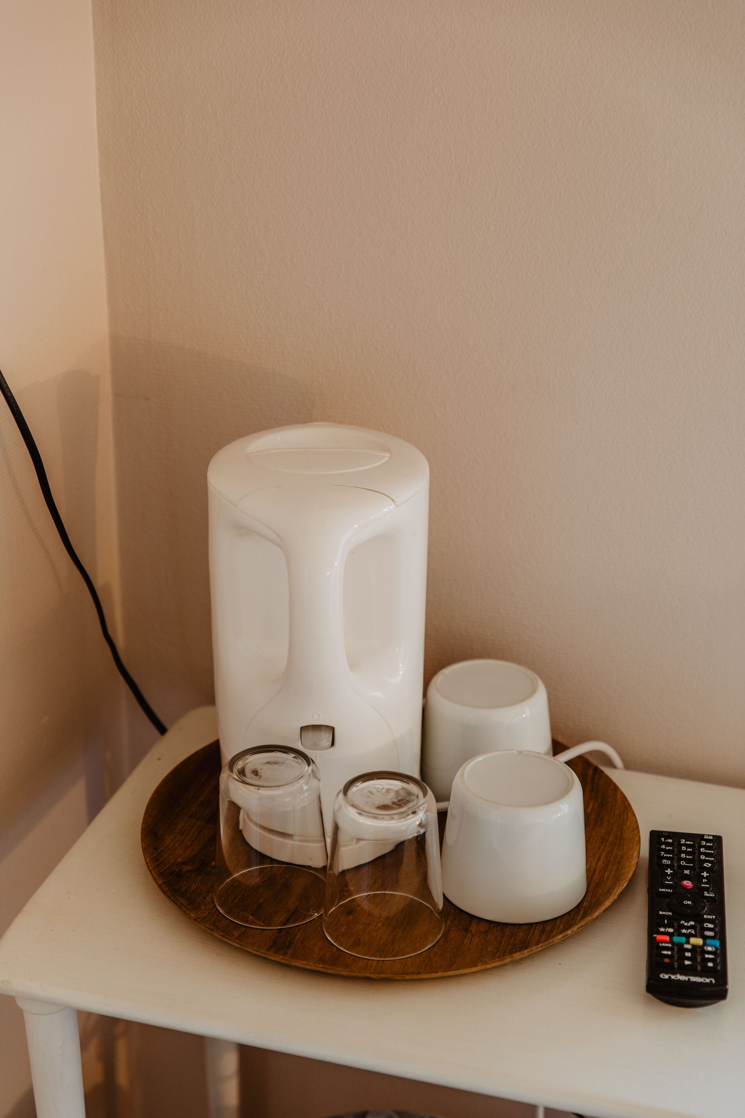 A white coffee maker, two white cups, and a remote control on a white table with a brown tray.