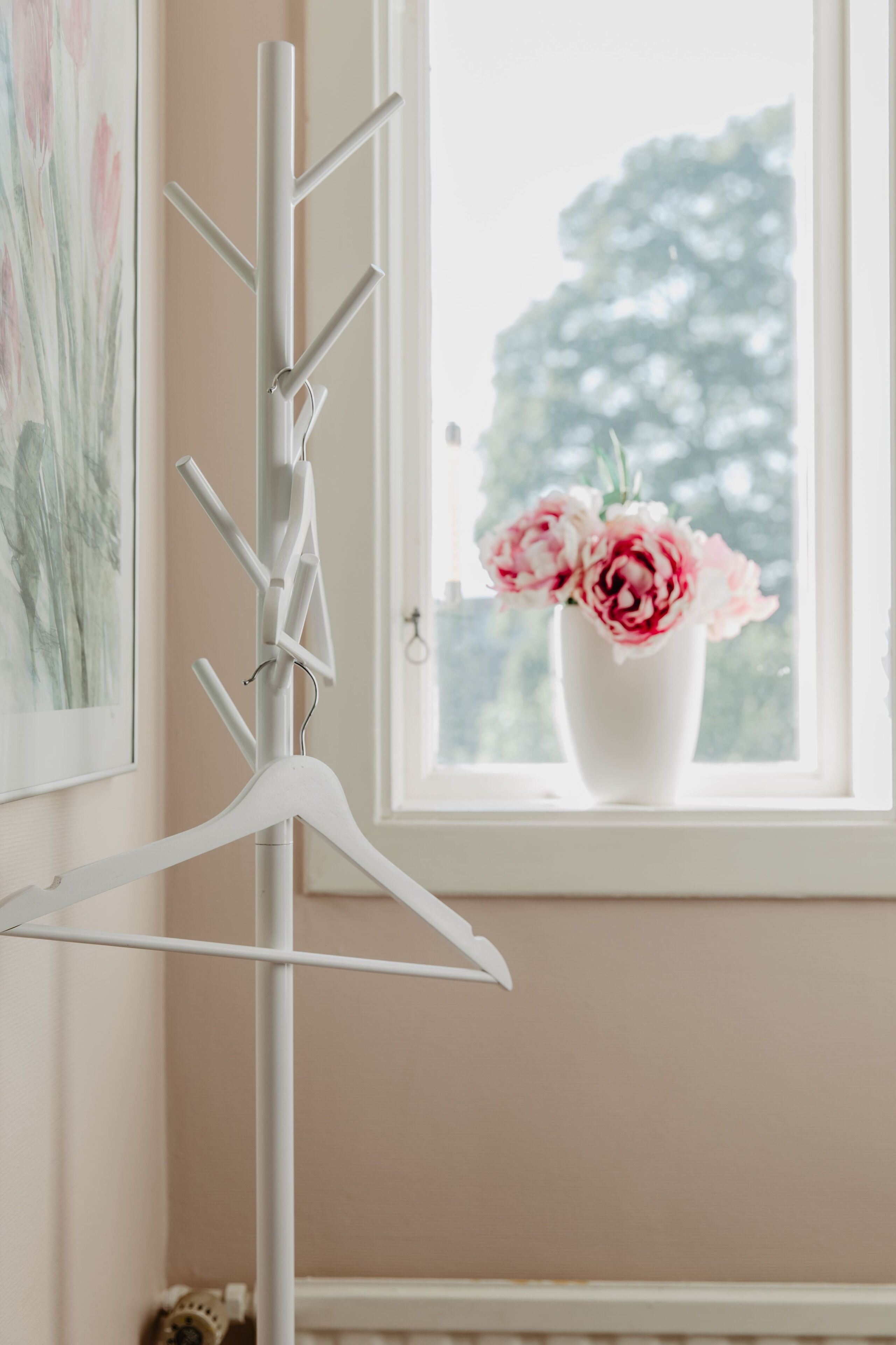 A room with a white coat rack, a framed floral artwork on the wall, and a vase with pink flowers on the windowsill.