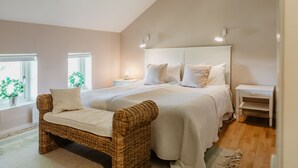 Un dormitorio con una cama grande, una silla de mimbre, una mesita de noche y un ventanal con plantas.