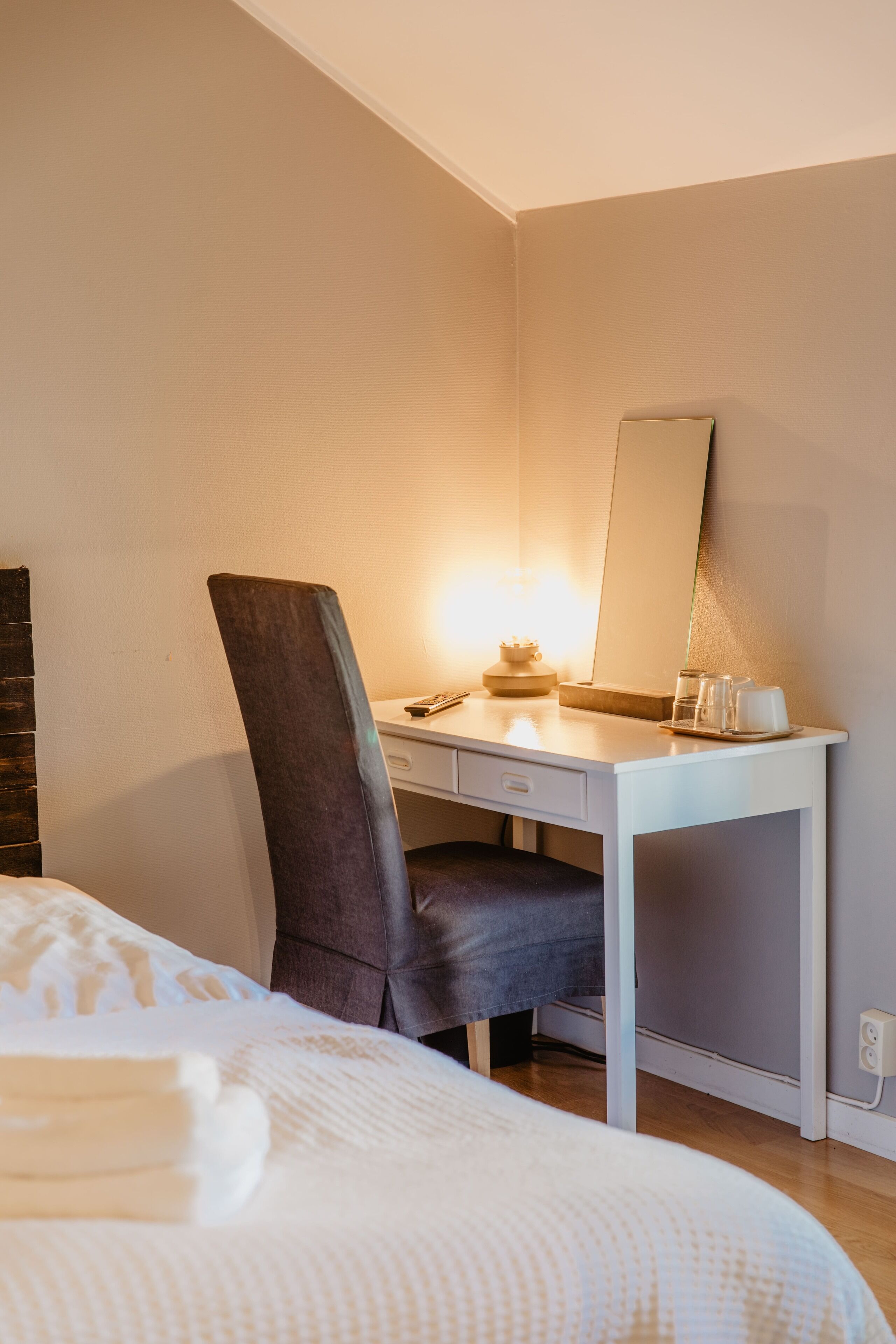 Une chambre d’hôtel comprenant un lit, un bureau avec une chaise, un miroir et une table de chevet avec une lampe et une tasse.