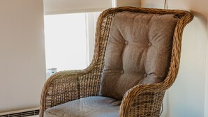 Un sillón de mimbre con un cojín gris en una habitación con un ventanal grande, un televisor de pantalla plana montado en la pared y una cama blanca con una colcha blanca.