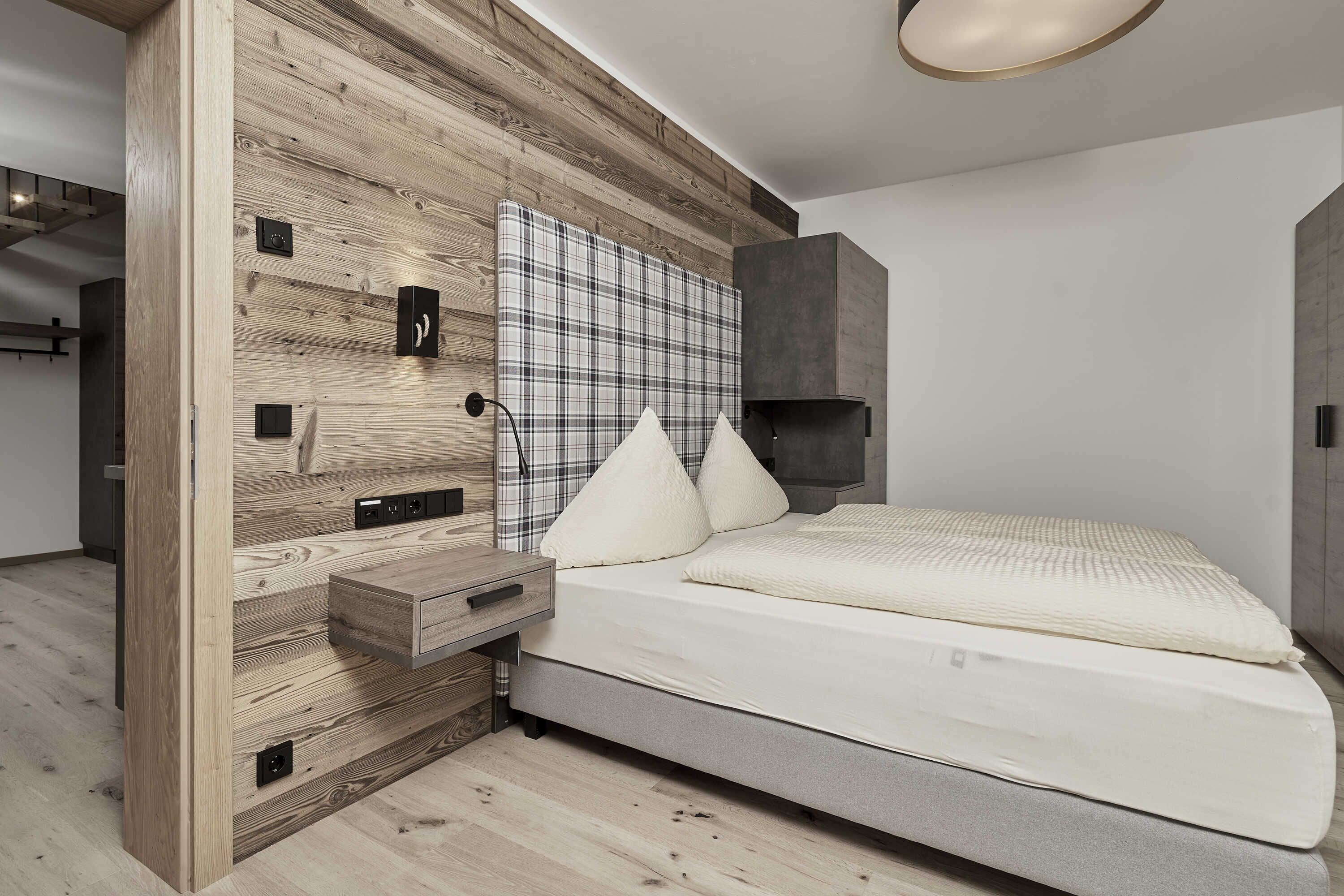 A modern bedroom with a wooden headboard, a large plaid-patterned wall hanging, a bed with white bedding, and a bedside table with a drawer.