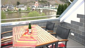 A balcony with a table, chairs, and a bottle of wine. A river and a bridge with a tower are visible in the background. - Ferienwohnungen Cella (Zell (Mosel))