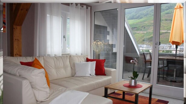 A modern living room with a white sofa, red and white cushions, a small wooden coffee table, and a view of a balcony with a glass door. - Ferienwohnungen Cella (Zell (Mosel))