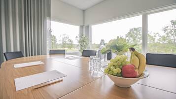 A conference room with a wooden table, meeting materials, a bowl of fruit, and large windows with a view of greenery.
