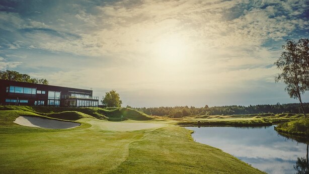 Ein modernes Gebäude mit großen Fenstern, ein gepflegter Golfplatz mit Teich und ein klarer Himmel.