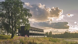 Ein modernes Gebäude mit flachem Dach und großen Fenstern, umgeben von einer grünen Fläche mit Wildblumen und einem Gew ässer im Hintergrund.