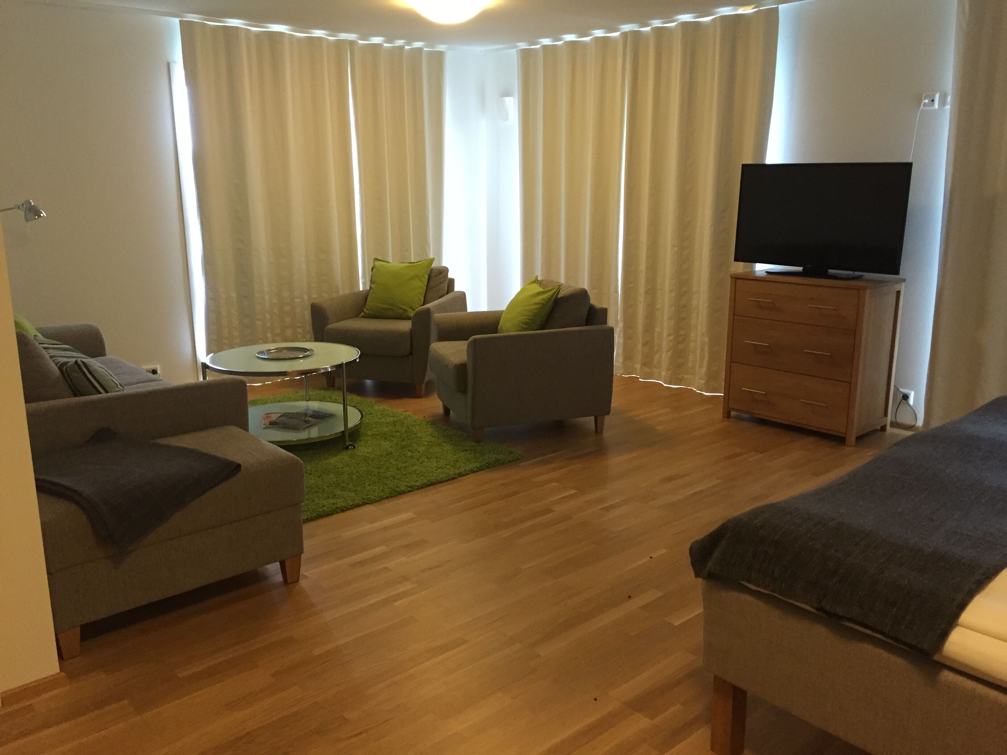 A modern living room with a sofa, two armchairs, a coffee table, a TV on a wooden cabinet, and a large window with curtains.