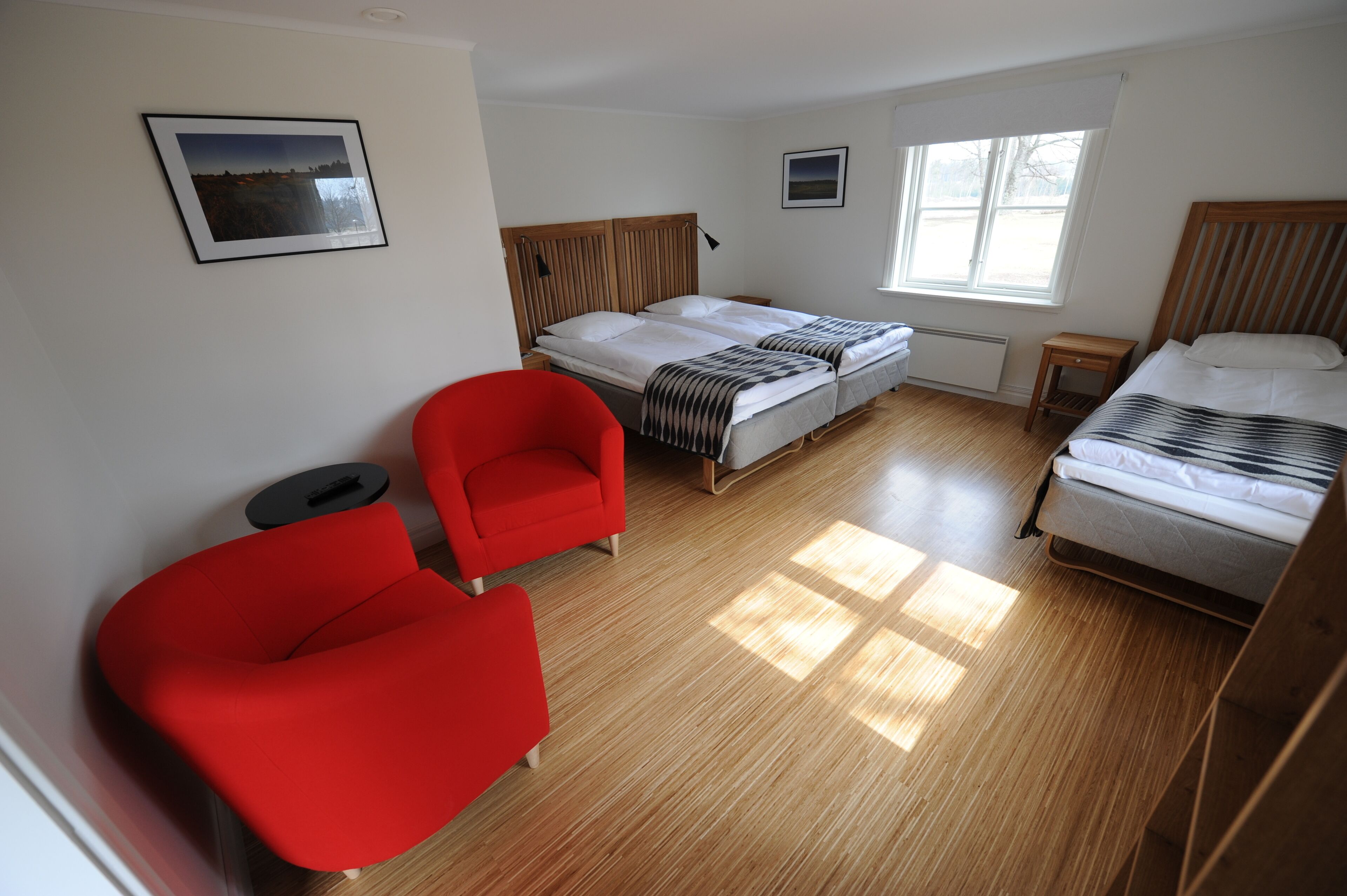 Habitación de hotel con dos camas, un sillón rojo, una mesita pequeña y una ventana con luz natural.