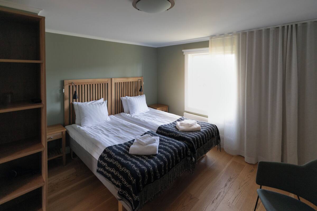 A hotel room with two beds, a wooden headboard, a nightstand, a chair, and a large window with curtains.