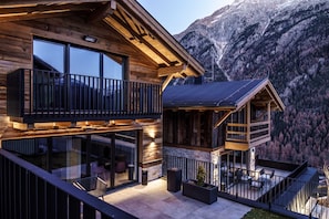 A modern wooden cabin with a balcony, large windows, and a view of a mountainous landscape.
