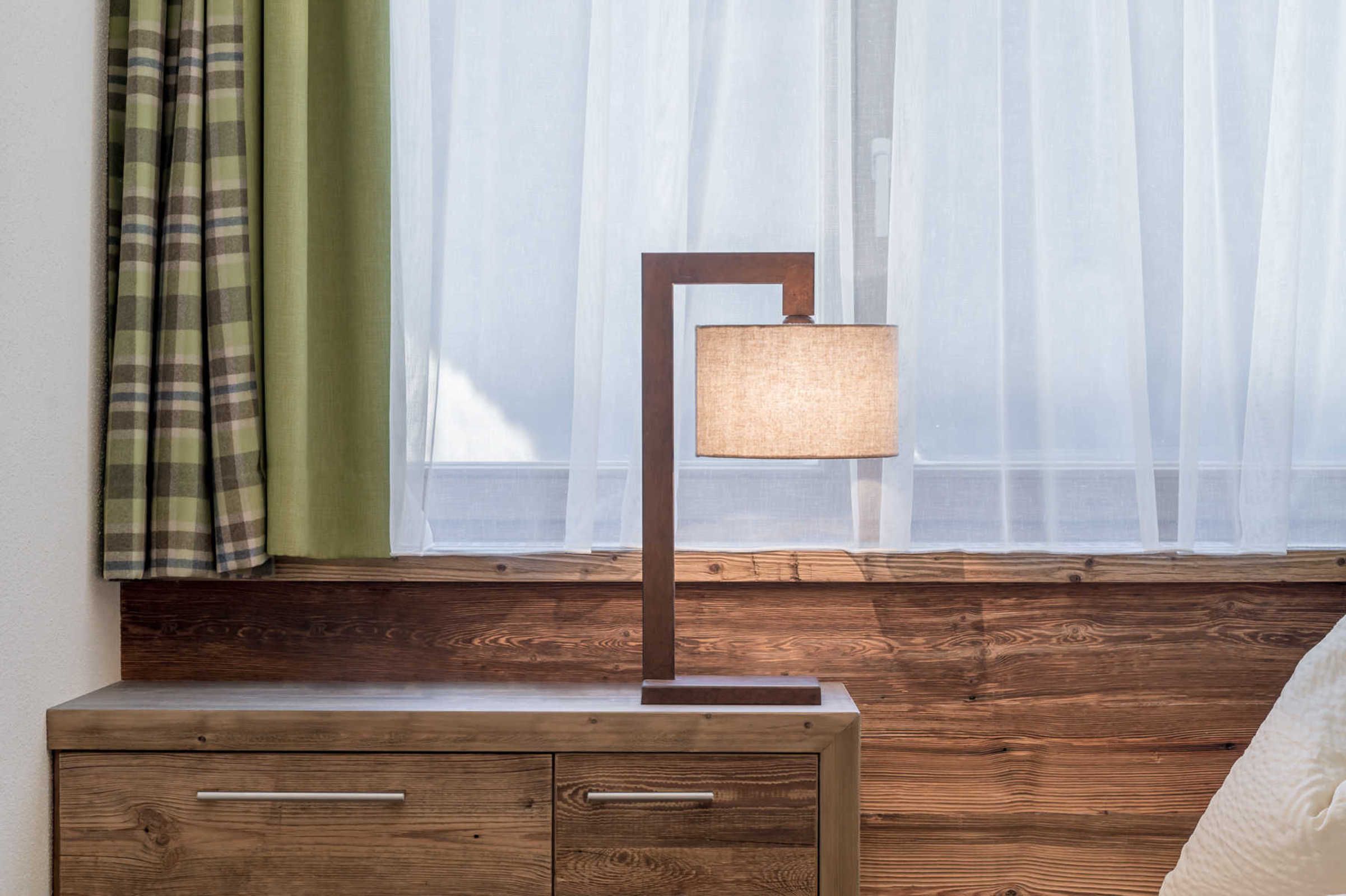 A wooden bedside table with a lamp, a window with sheer curtains, and a plaid curtain.