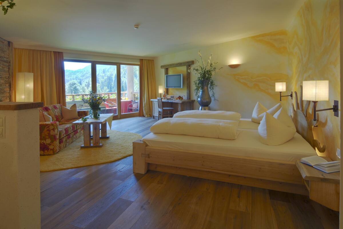 Habitación de hotel con una cama grande, un escritorio con ordenador, televisión y balcón con vistas a las montañas.