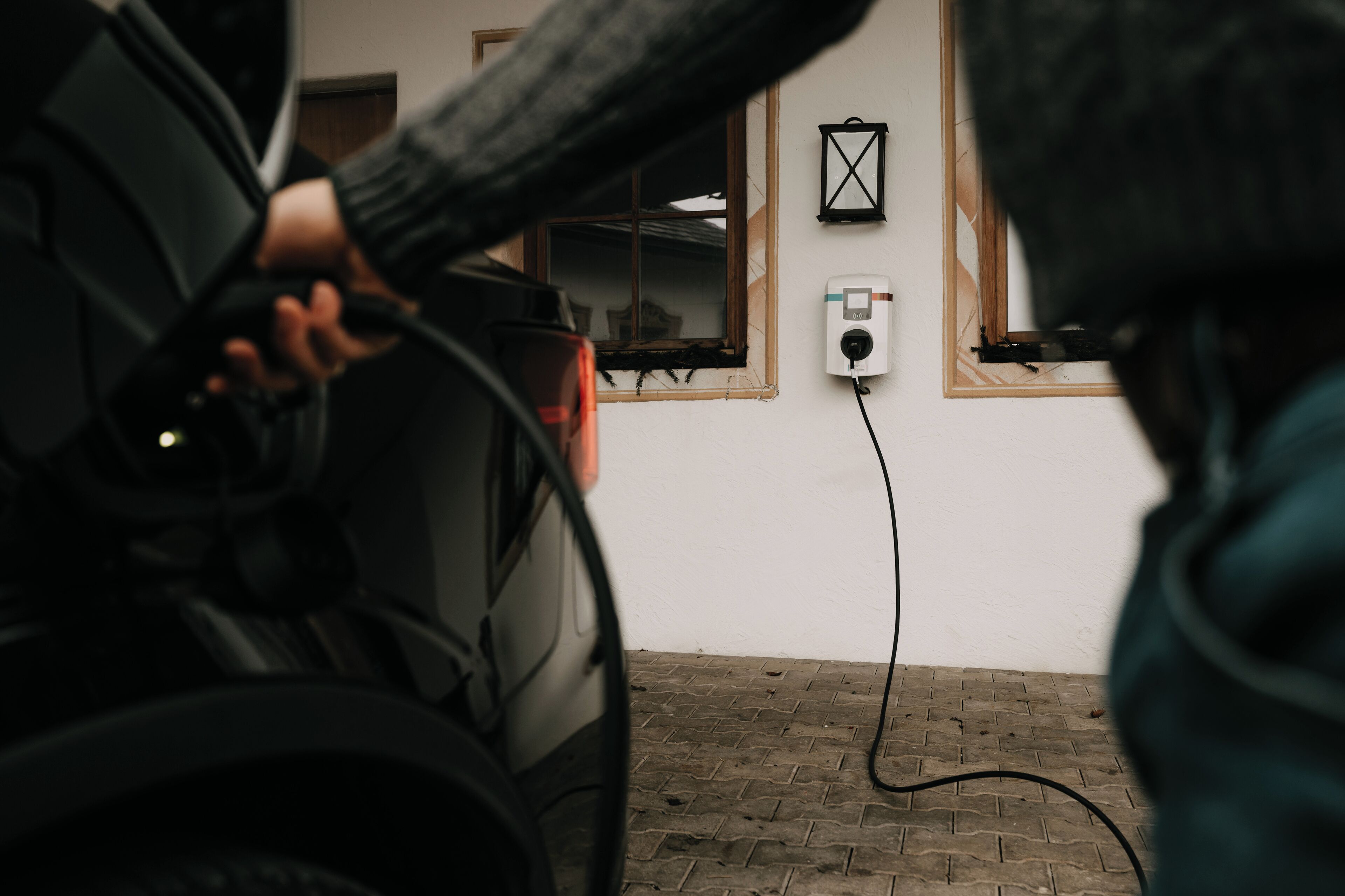 A person charging a device at a wall socket in a room with a brick floor and wooden trim around the windows.