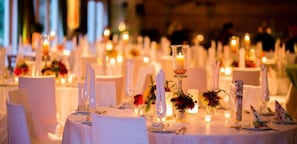 Ein Festsaal mit runden Tischen, gedeckt für eine festliche Veranstaltung, mit weißen Tischdecken, Kerzen und floralen Tischarrangements.
