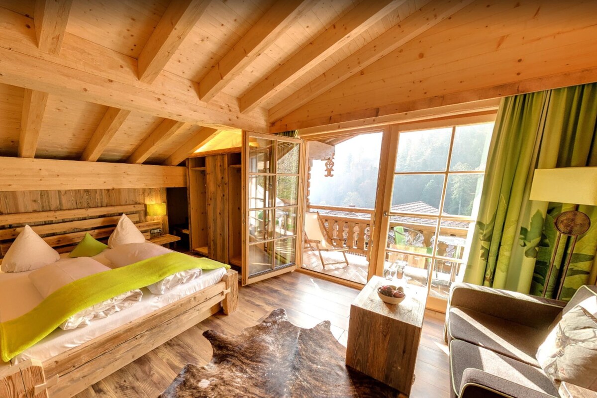 Ein Schlafzimmer in einer Holzhütte mit Bett, Sofa, einem kleinen Tisch und Blick ins Freie.