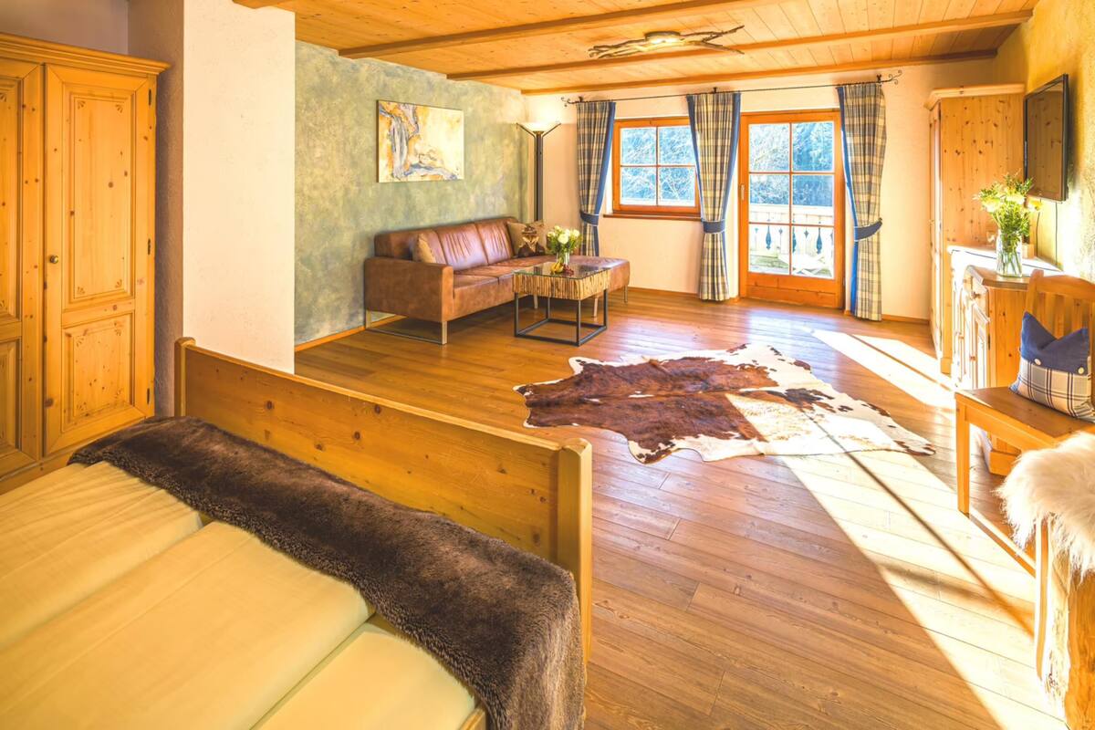 Una habitación amplia con piso de madera, un ventanal con cortinas, un sofá, una mesa de centro y una cama con una manta de piel.