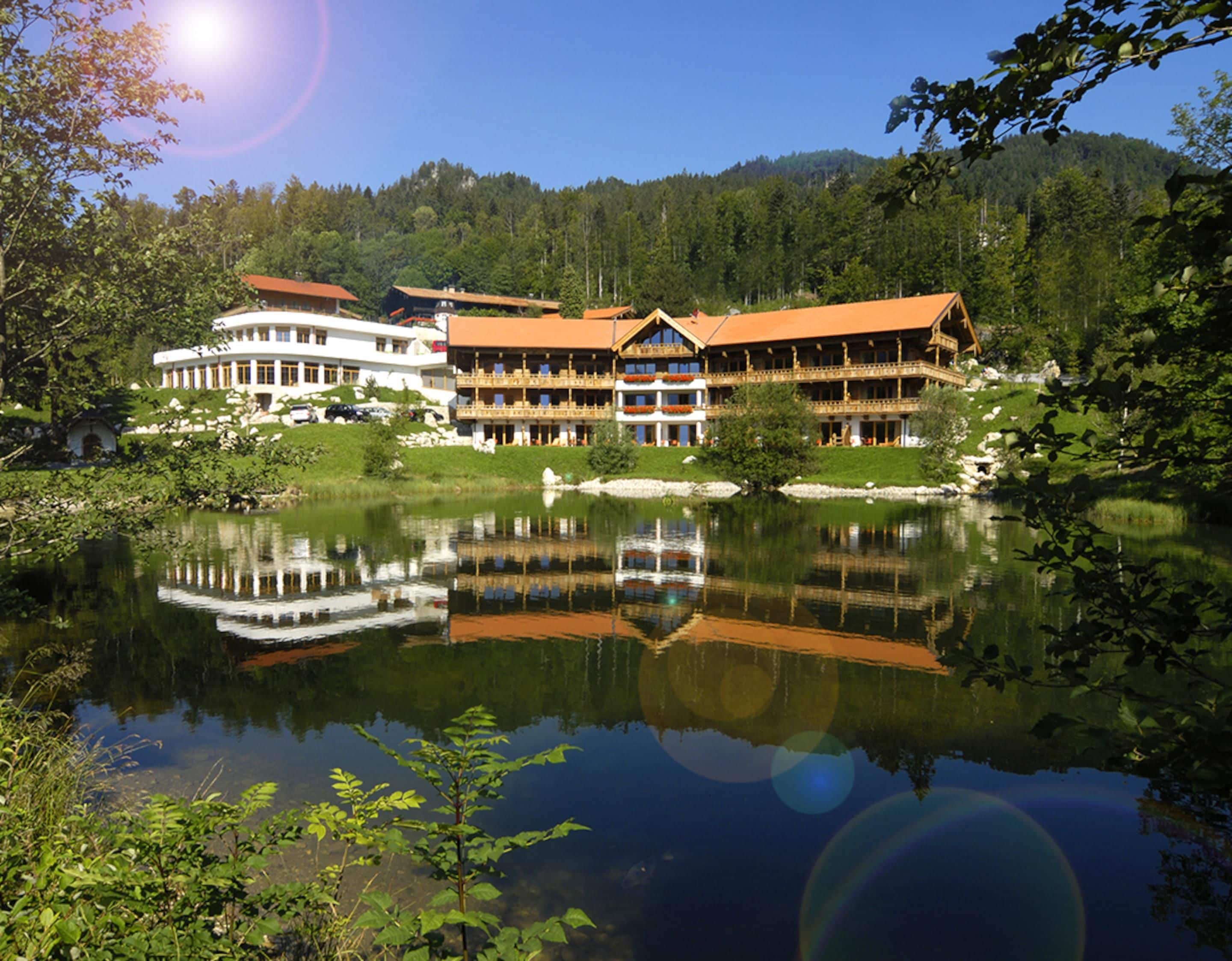 A large, multi-story hotel with a reflection in a lake, surrounded by greenery and hills.