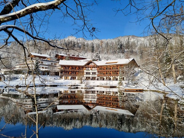 Ein großes, mehrstöckiges Holzhaus mit Balkonen, gelegen an einem ruhigen See, umgeben von verschneiten Landschaften.