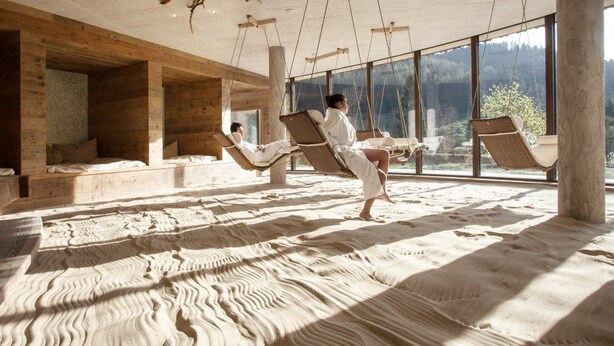 Una habitación con paredes de madera y ventanas amplias, una cama y dos personas relajándose en columpios.
