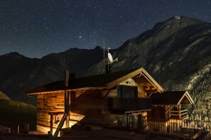 A wooden cabin with a satellite dish, surrounded by mountains under a starry night sky.