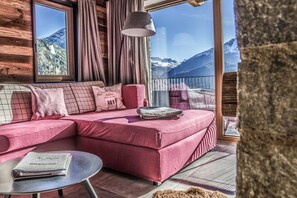 Une pièce chaleureuse avec un canapé rouge, une petite table sur laquelle repose un livre, et une vue sur les montagnes par de grandes fenêtres.