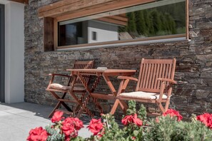 A patio with a table and chairs, a stone wall, and a window overlooking greenery.