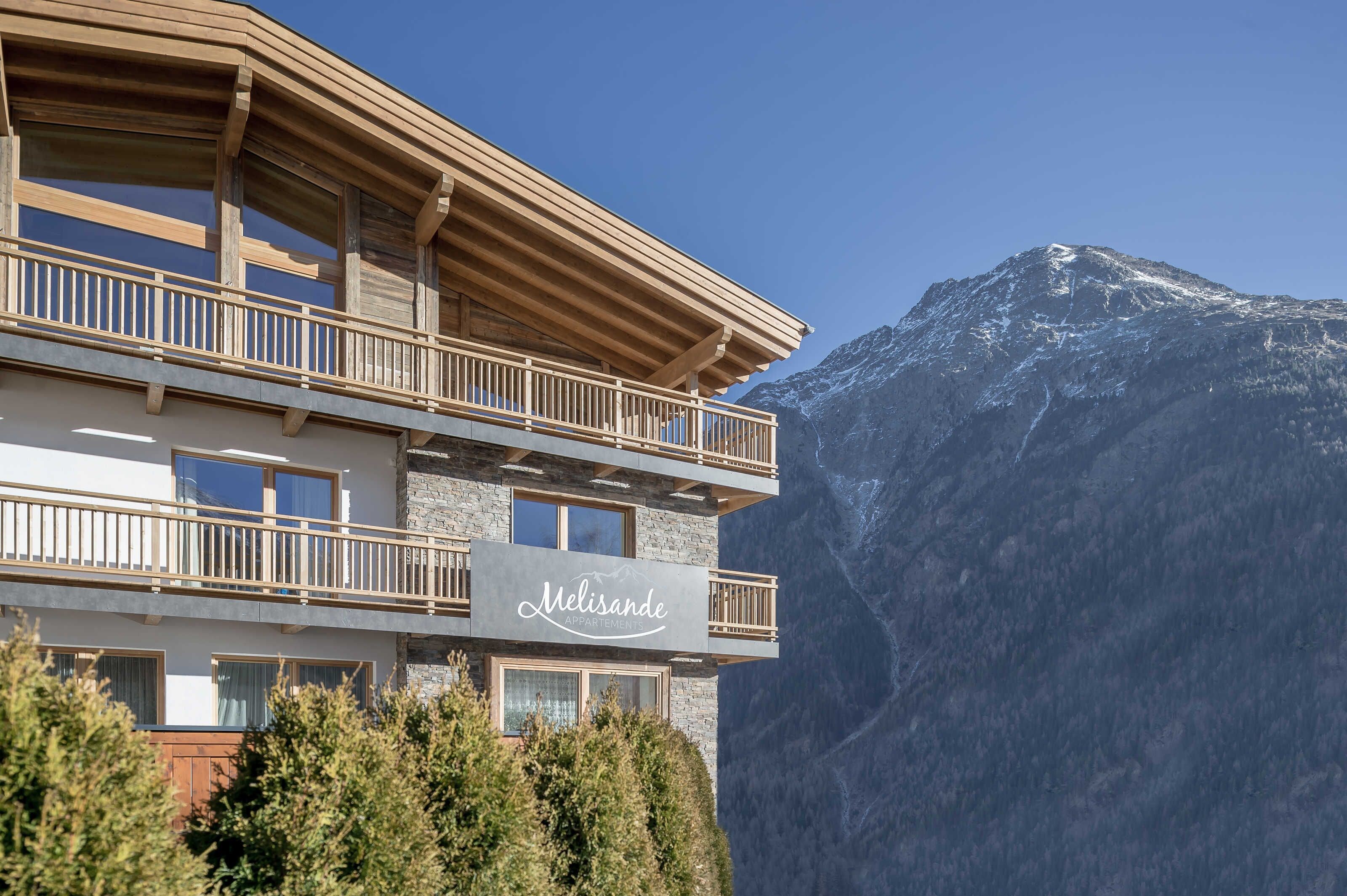 A multi-story building with balconies, a sign reading 'Melisande Apartments,' and a mountainous backdrop.